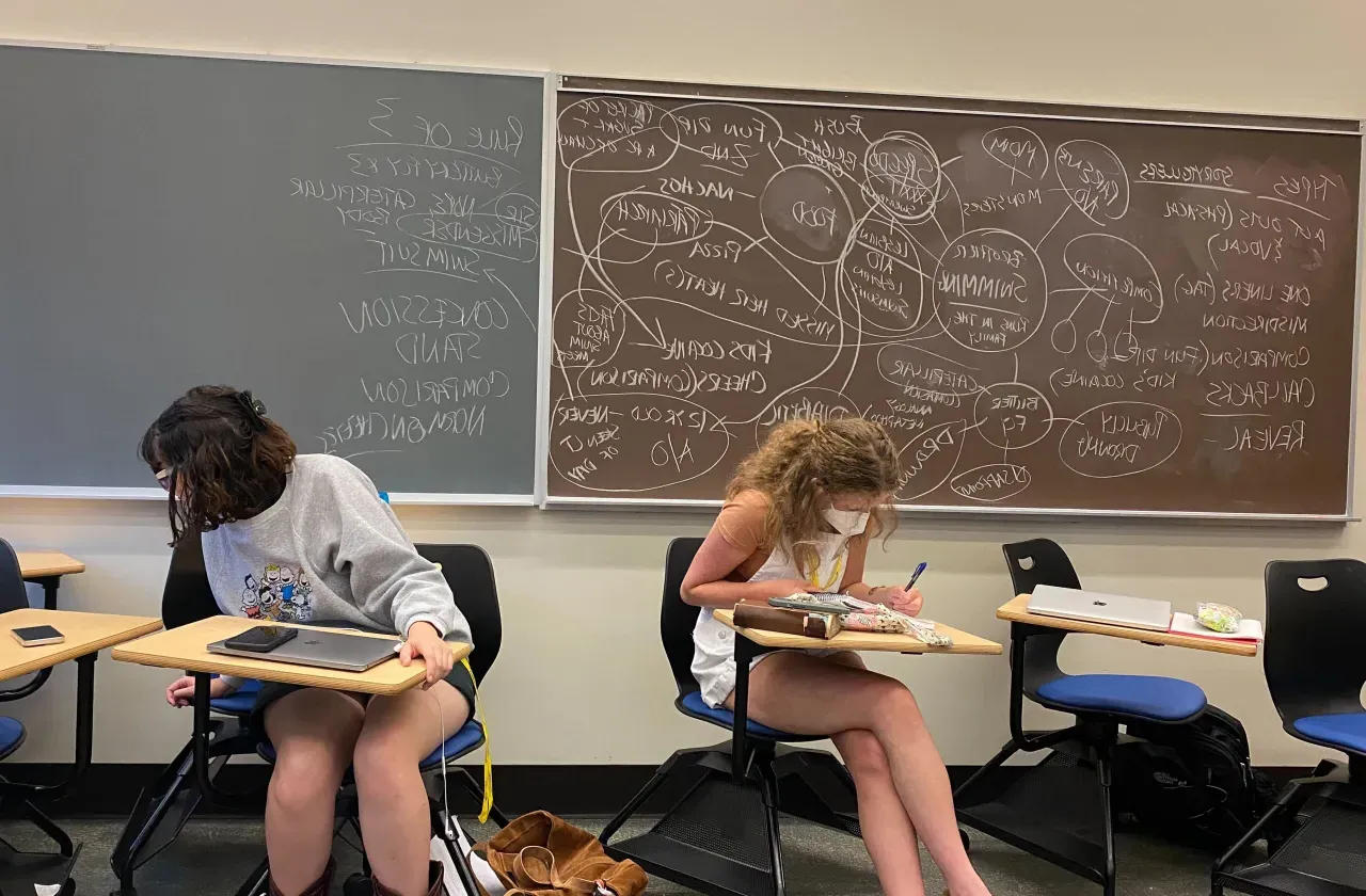 Two students writing, with a filled chalkboard behind them.