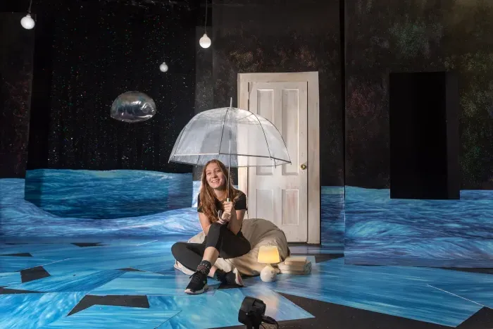 A student sitting on a stage holding an umbrella over her head.